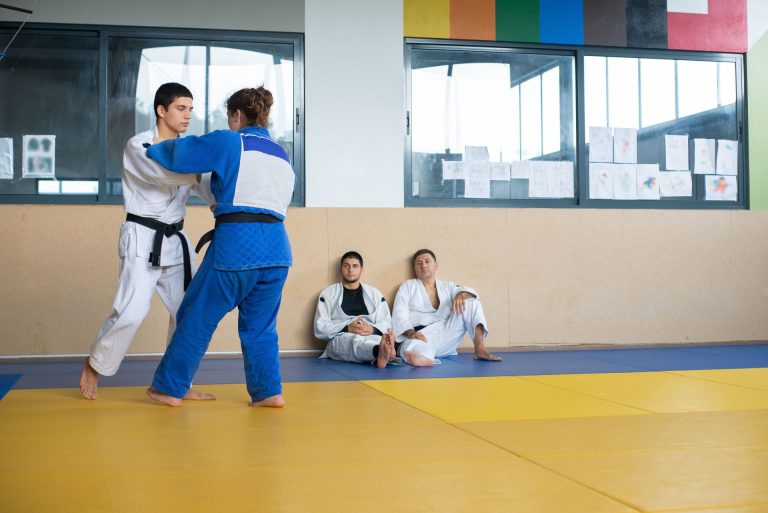 Male and female judokas practice judo techniques on dojo mat in an indoor setting, observed by fellow trainees.